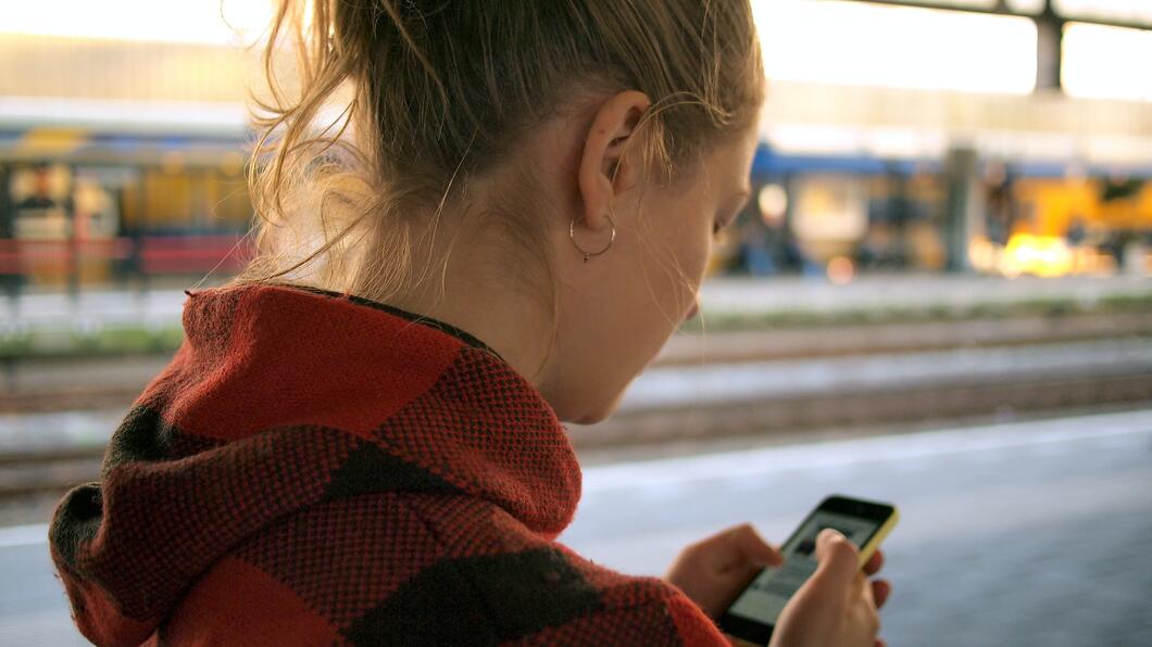Persoon staat met telefoon in de hand. Ze draagt een rode trui en staat op een station.