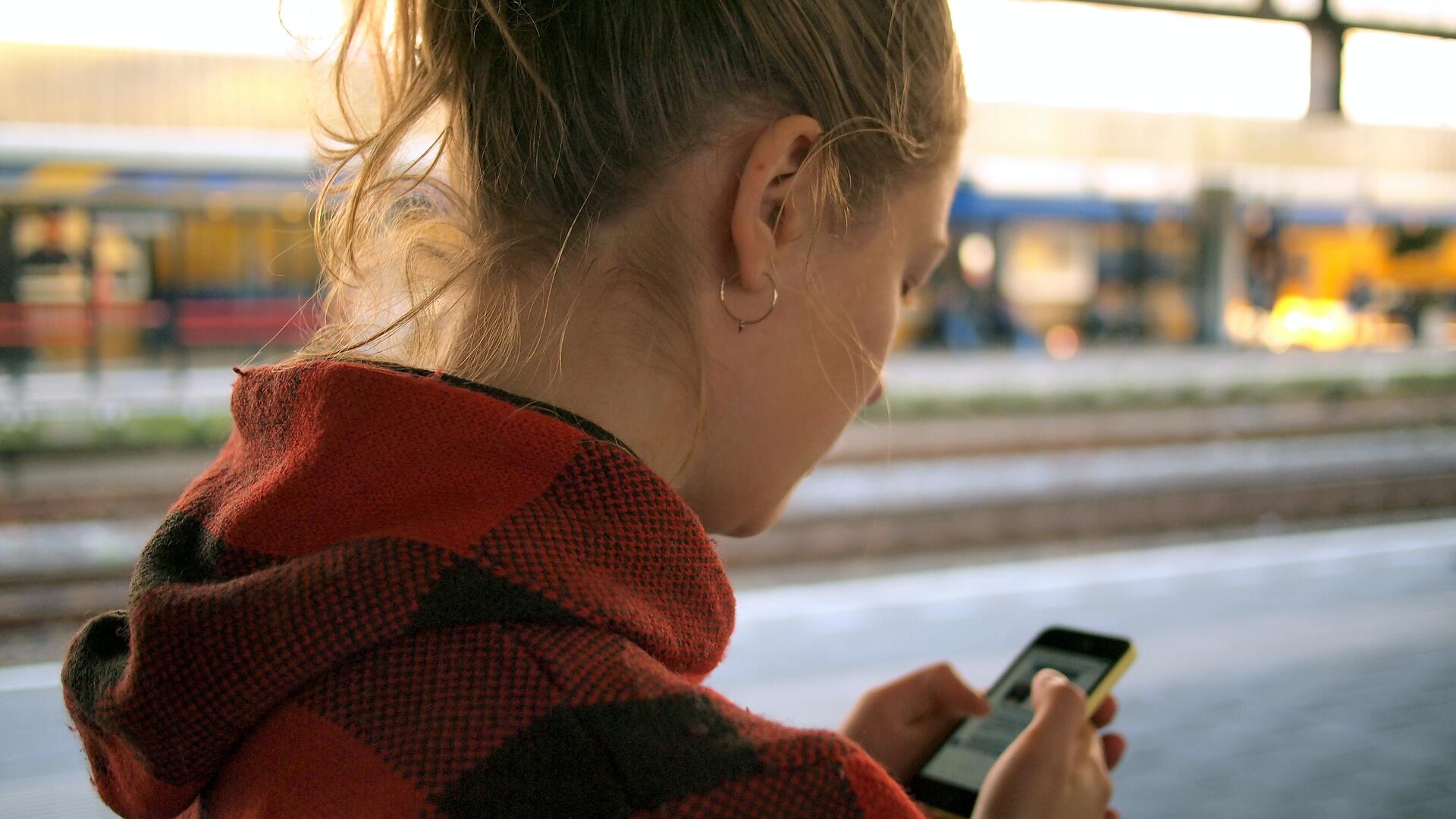 Persoon staat met telefoon in de hand. Ze draagt een rode trui en staat op een station. 