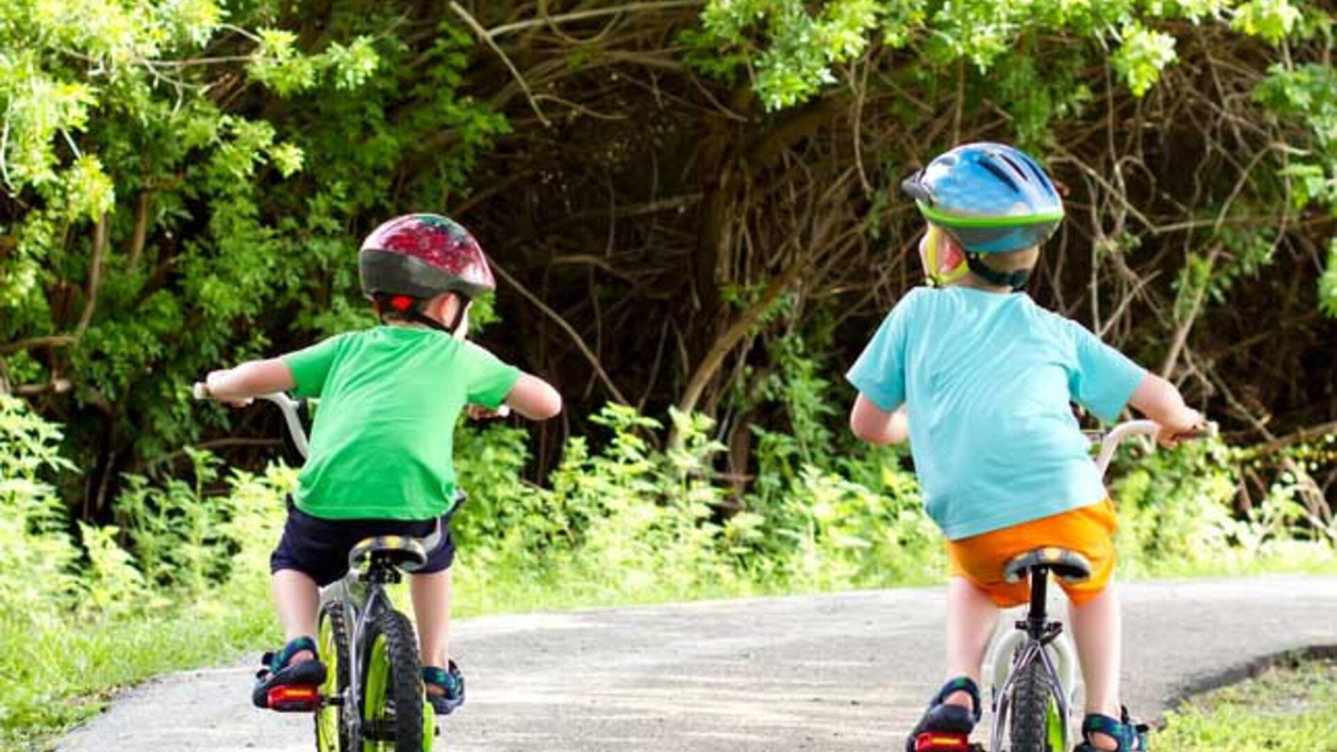 Twee kindjes op fietsjes met zijwieltjes met groene struiken op de achtergrond