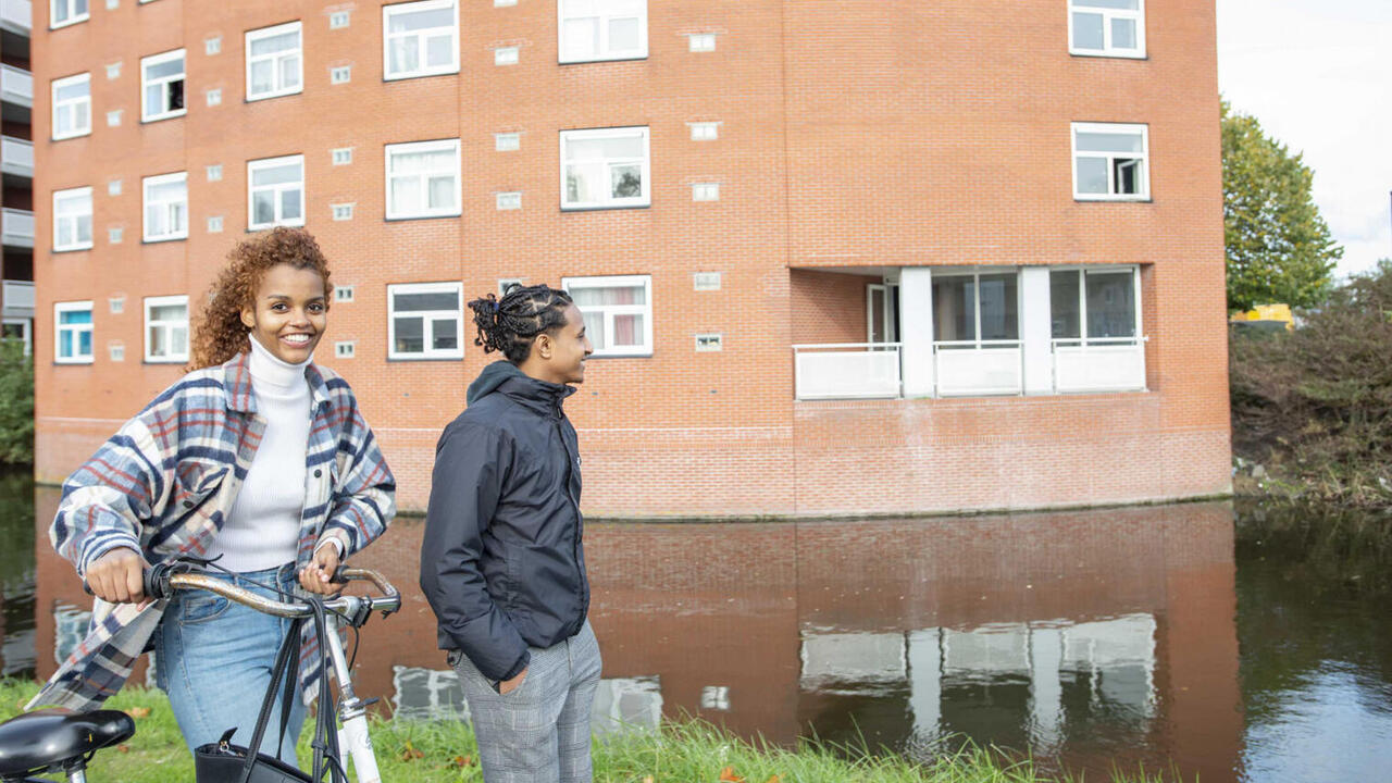 Twee jongeren lopen langs woningen en water. Een heeft een fiets bij zich.