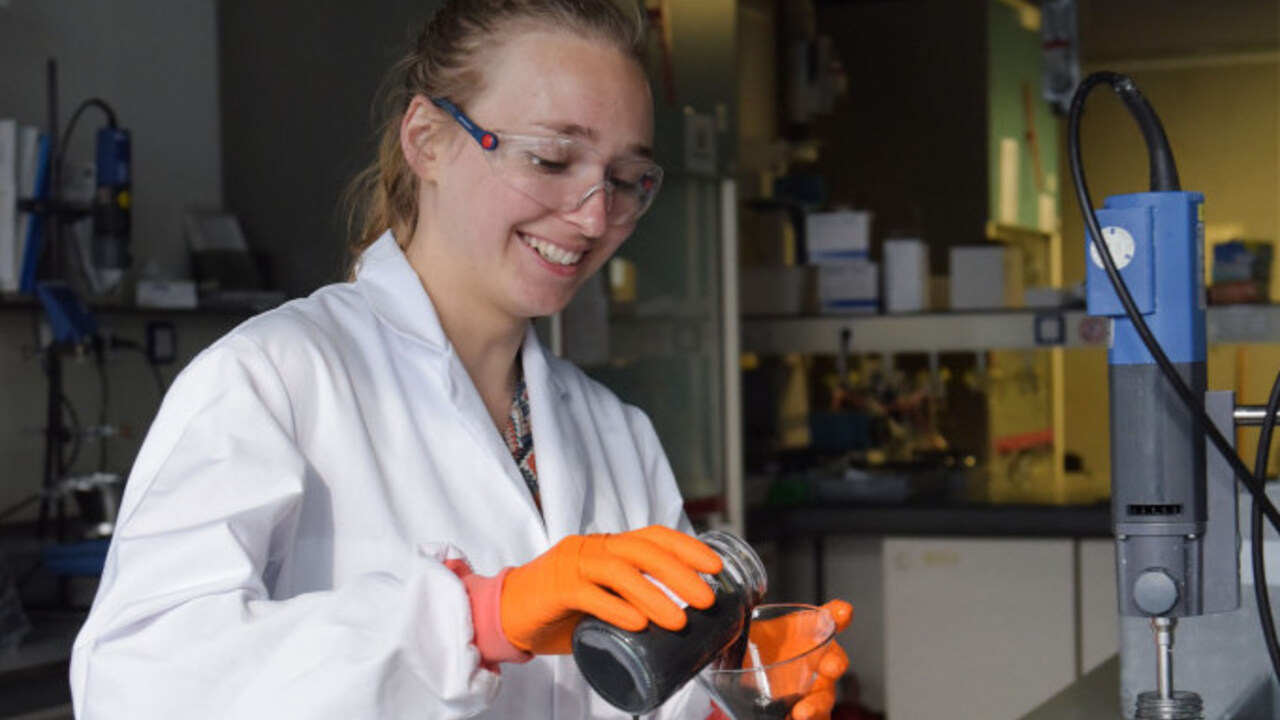 Vrouw in labjas en veiligheidsbril giet iets zwarts uit een pot in een glazen trechter.